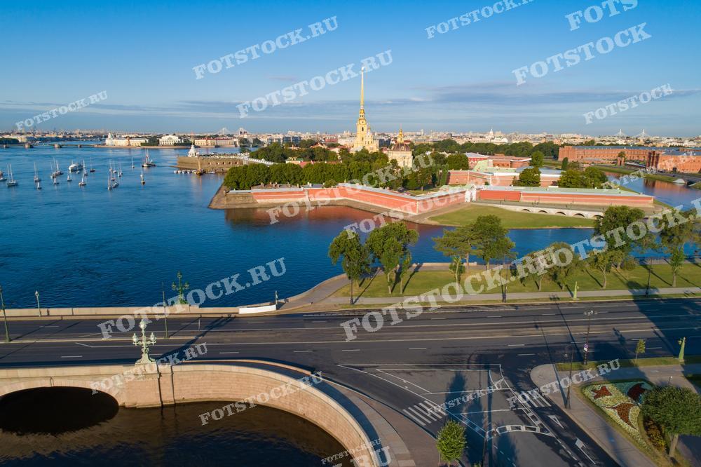 На фотографии изображена Петропавловская крепость в Санкт-Петербурге, окруженная рекой Невой. Видна золотая игла Петропавловского собора, возвышающаяся над крепостью. Спокойная утренняя река отражает ясное небо, на воде видны яхты и катера. Зеленые деревья и газоны добавляют свежести и контраста городскому пейзажу.